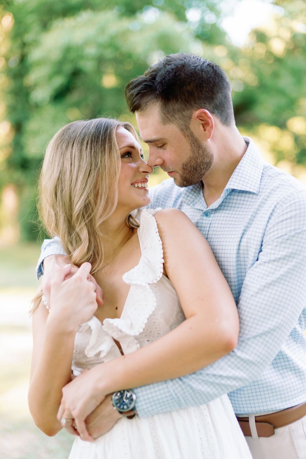 Couple nearly kissing in golden hour light, man embracing woman in white ruffled dress in a green garden.