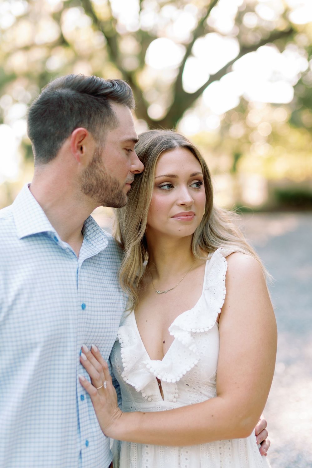 Close romantic portrait of an engaged couple outdoors, woman in white ruffled dress with ring visible.