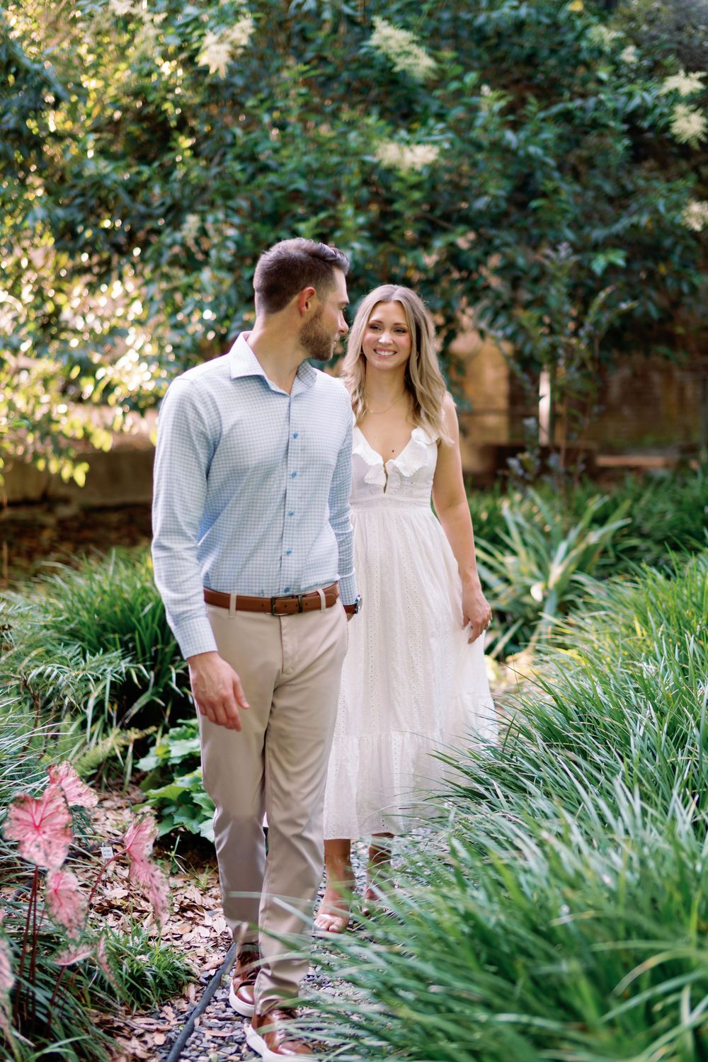 Couple walking hand-in-hand through a lush garden path, woman in white dress smiling at man in light blue shirt.