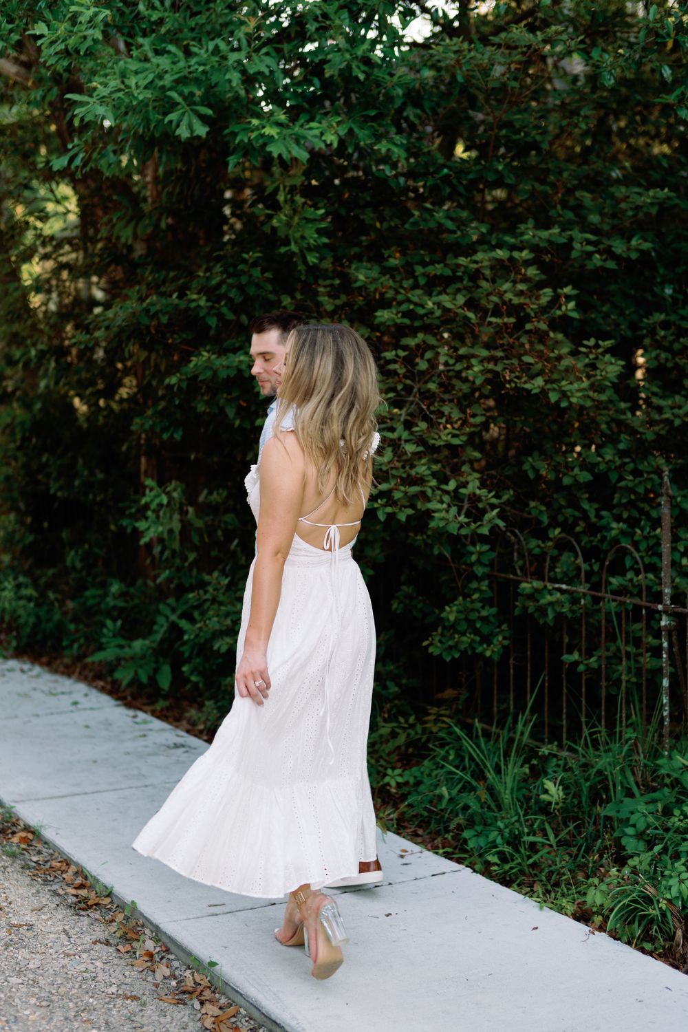 Woman in backless white midi dress and heels walking on garden path surrounded by dense green hedges.