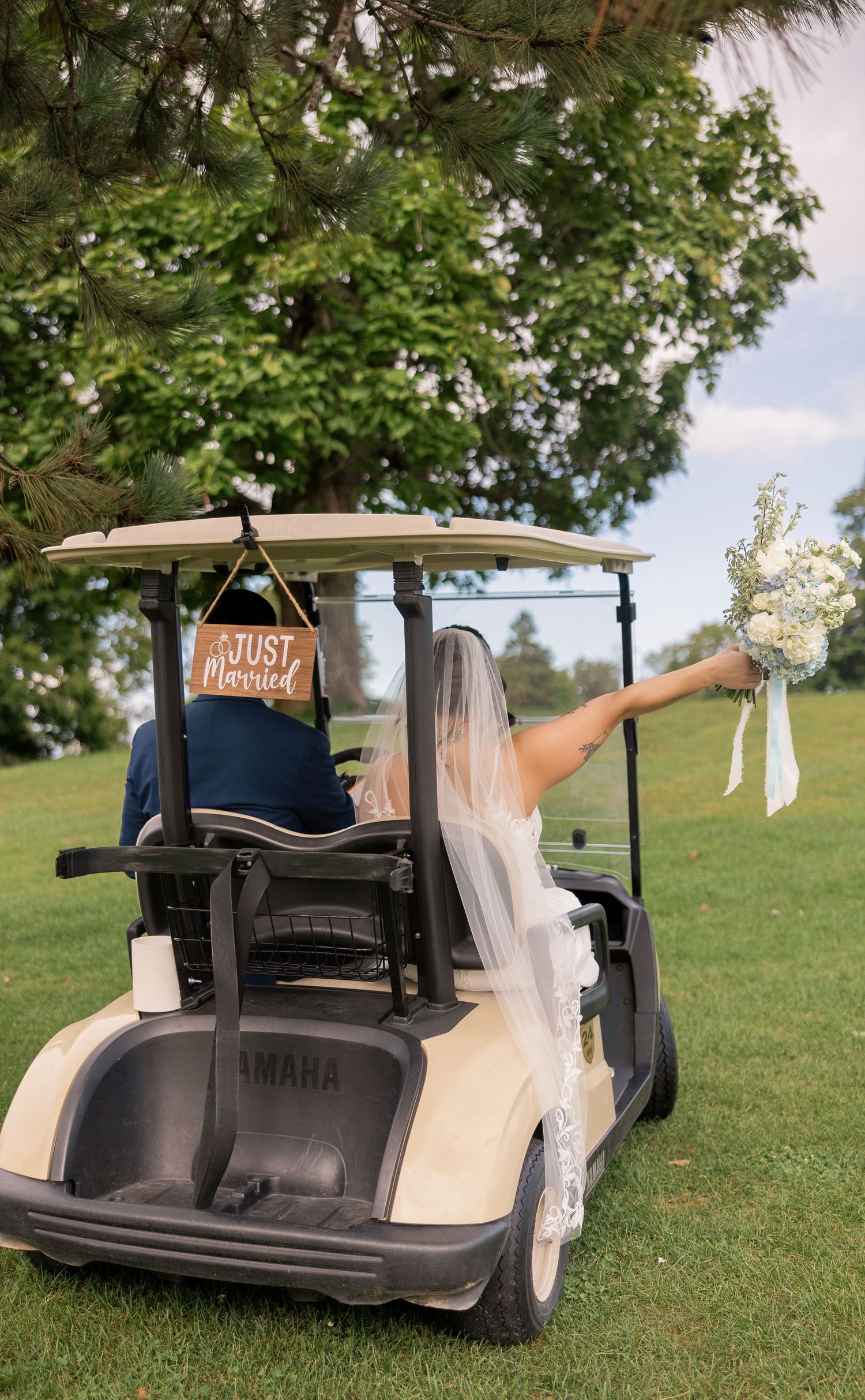 Newlyweds ride a Yamaha golf cart with a Just Married sign, bride holding bouquet aloft on a green golf course.