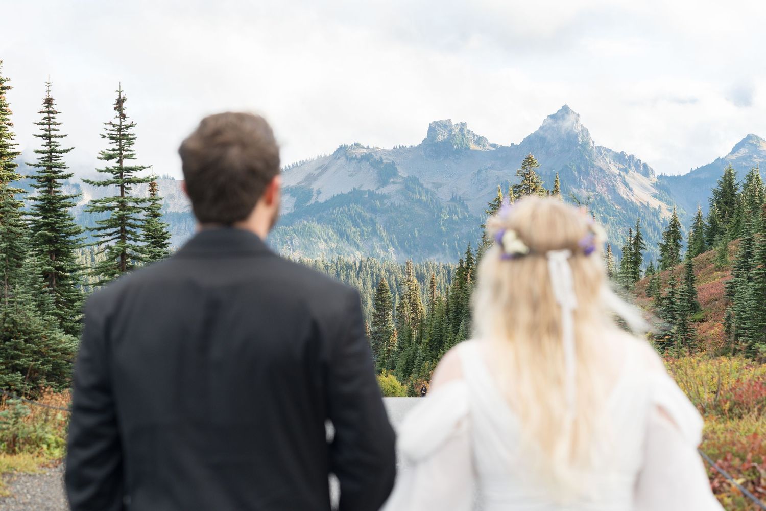 Bride in white gown standing on rocky lakeside shore with mountain forest backdrop on a sunny day.
