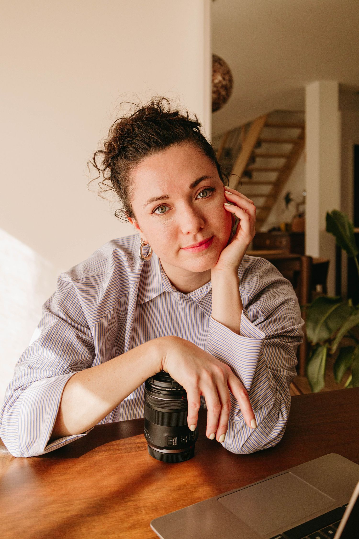 Woman with curly hair resting her chin on her hand at a wooden desk with a camera lens and laptop.