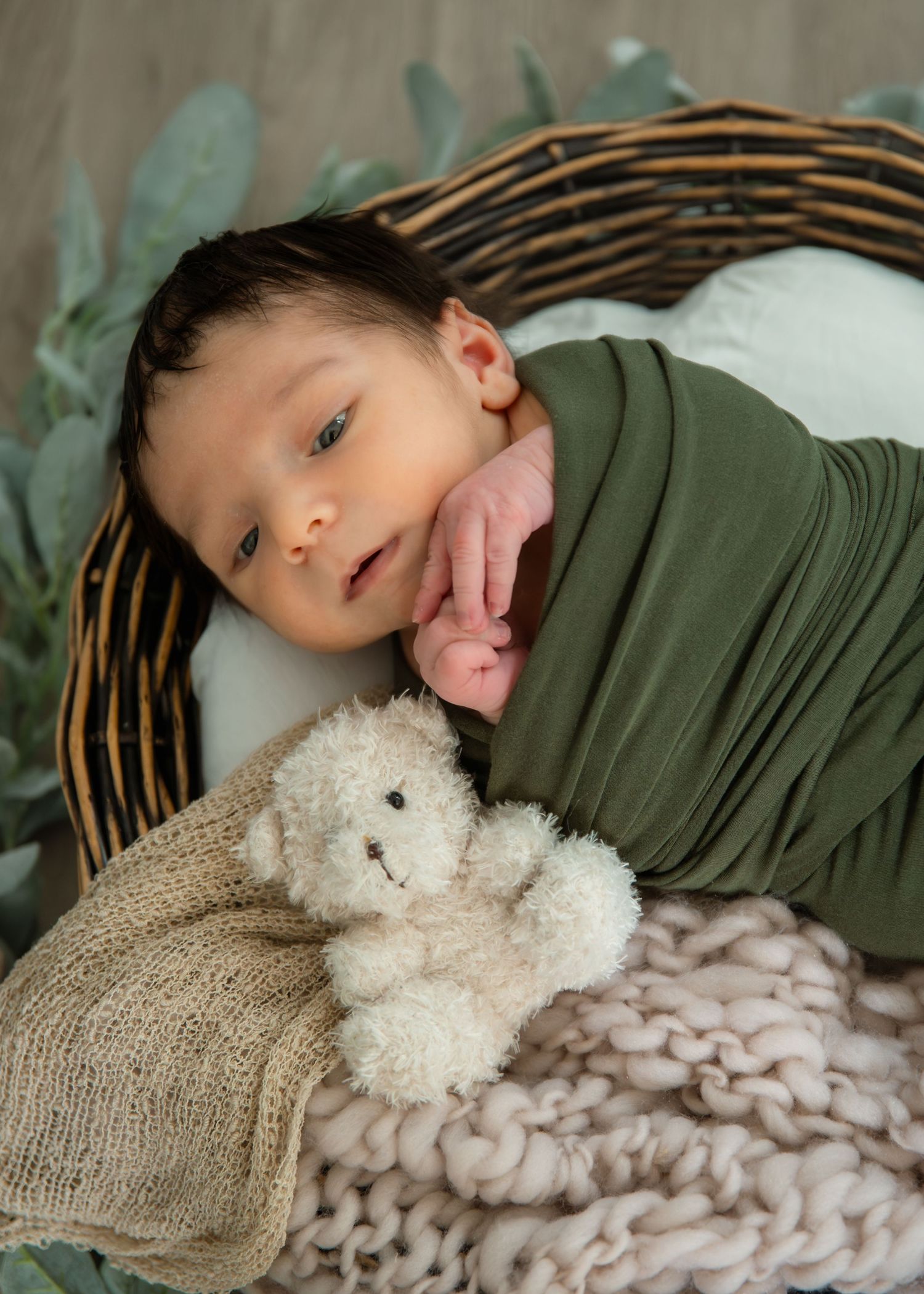 Newborn baby swaddled in green wrap resting in a wicker basket with a white teddy bear and pink knit blanket.
