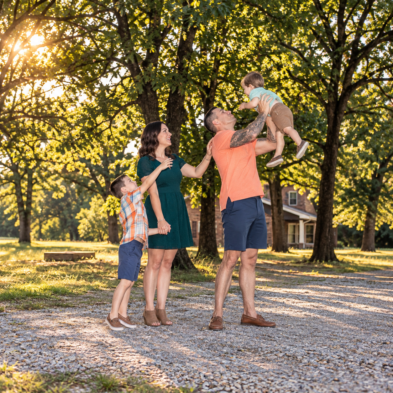 Family enjoying a sunny day outdoors, dad tossing baby in the air while mom and child watch, surrounded by lush green trees.