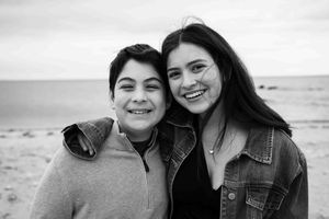 Brother and sister pose together on a beach in a black and white photo, one in a hoodie, one in a denim jacket.