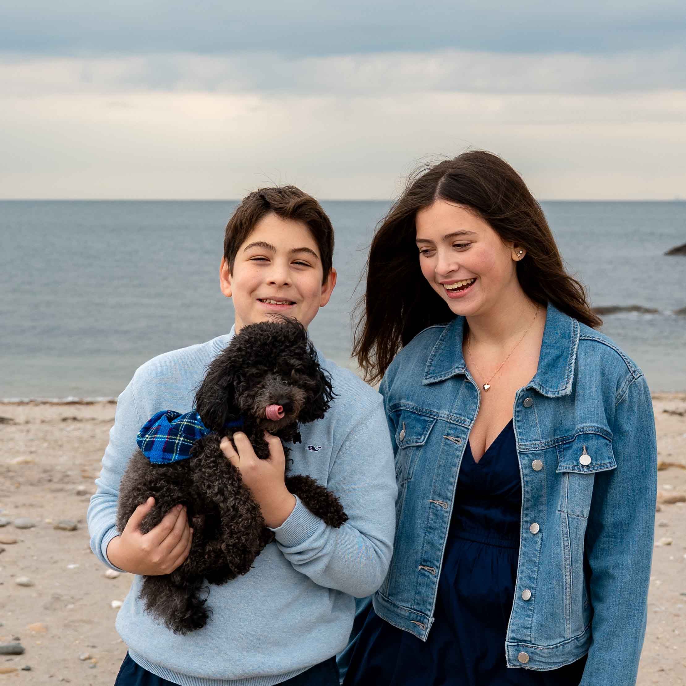 Two smiling teens with a black curly dog on a rocky beach with ocean backdrop.