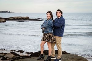 Teen siblings pose back-to-back on rocky coastal shoreline with ocean waves in background.