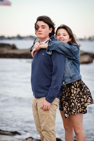 Teen siblings posing together on rocky beach at sunset, girl in denim jacket hugging boy in blue sweater.