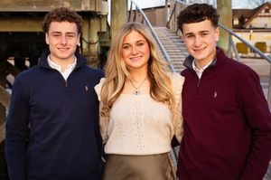 Three young adult siblings smiling together outdoors near a waterfront bridge on a sunny day.