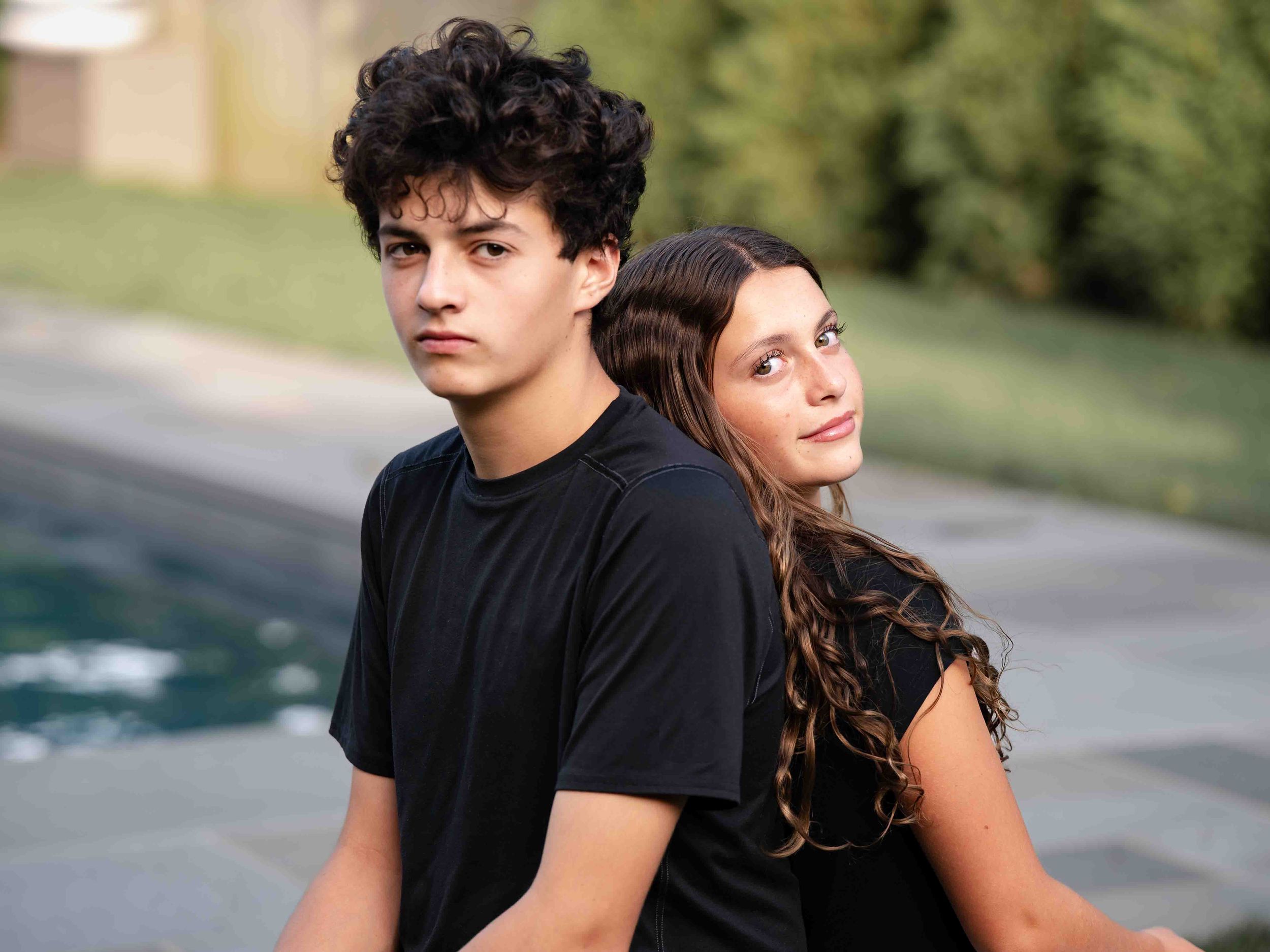 Two teenage siblings in black t-shirts pose back-to-back near a pool in an outdoor portrait photo.