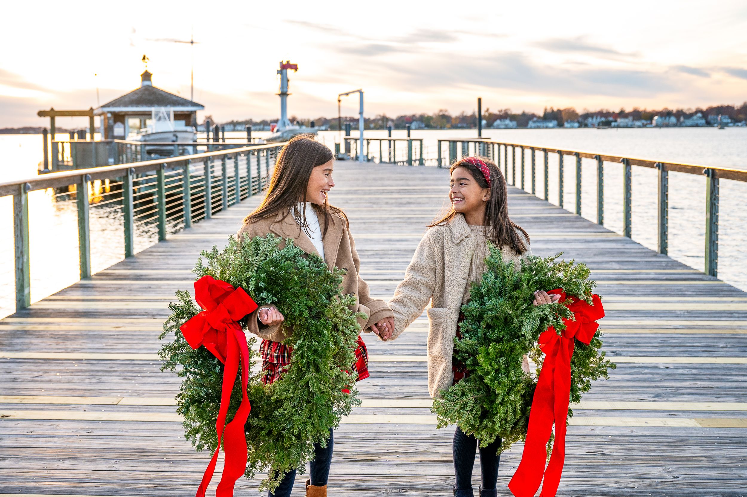 Sisters holding Christmas wreaths with red bows on a wooden pier by the water at sunset.