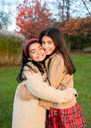 Two young girls hugging and smiling outdoors in autumn, wearing cozy coats near colorful fall foliage.
