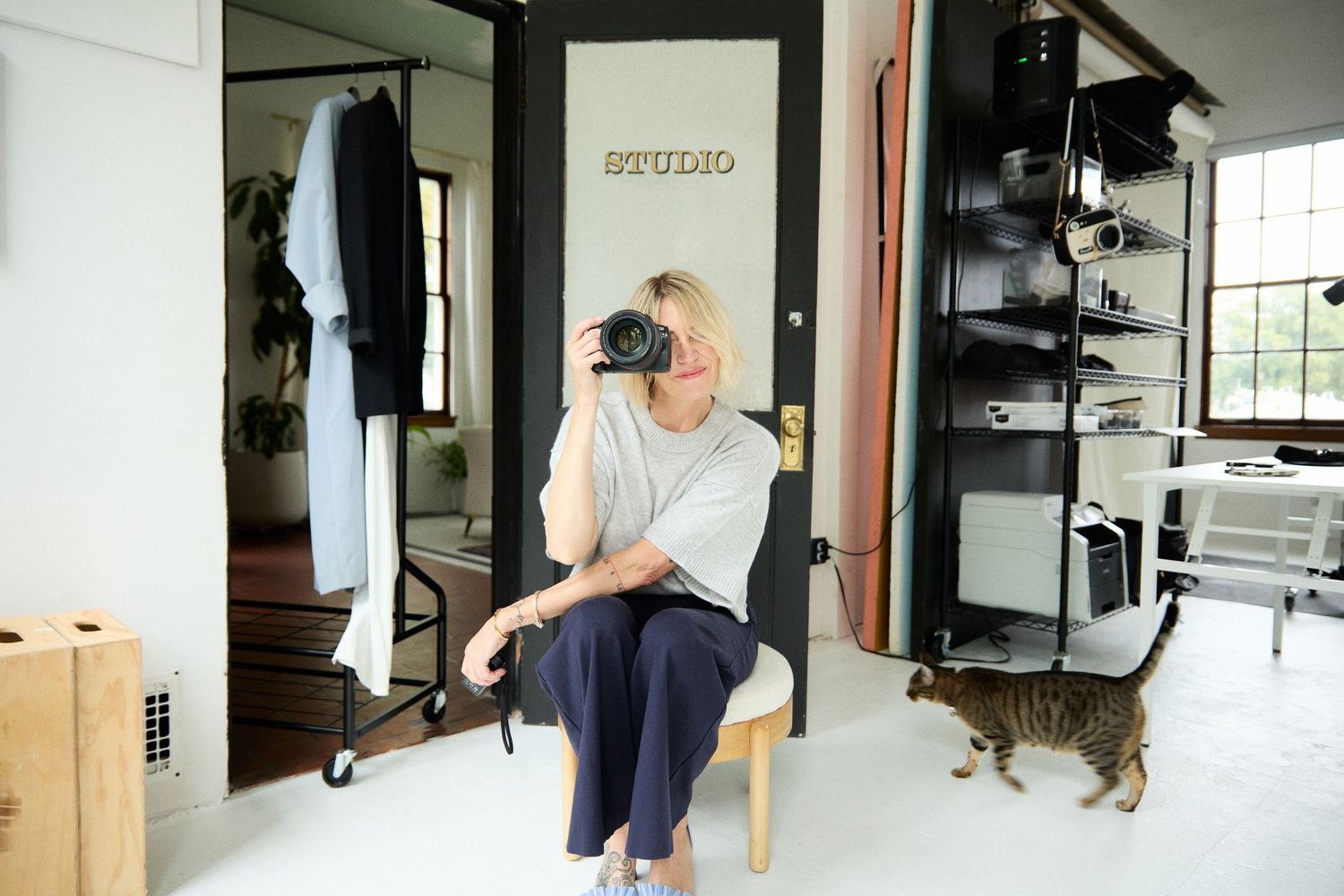 Photographer sitting on chair in bright studio holding camera, with tabby cat nearby and clothing rack beside mirror.