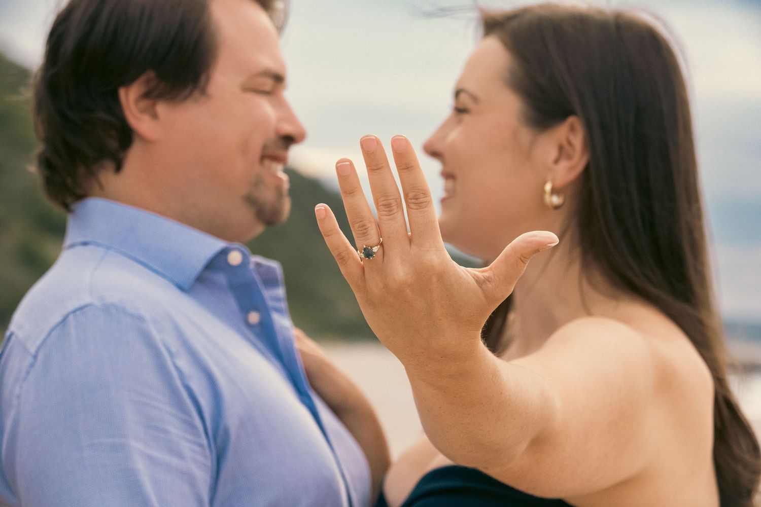Smiling woman shows off her sparkling engagement ring while a happy bearded man in a blue shirt stands close beside her.