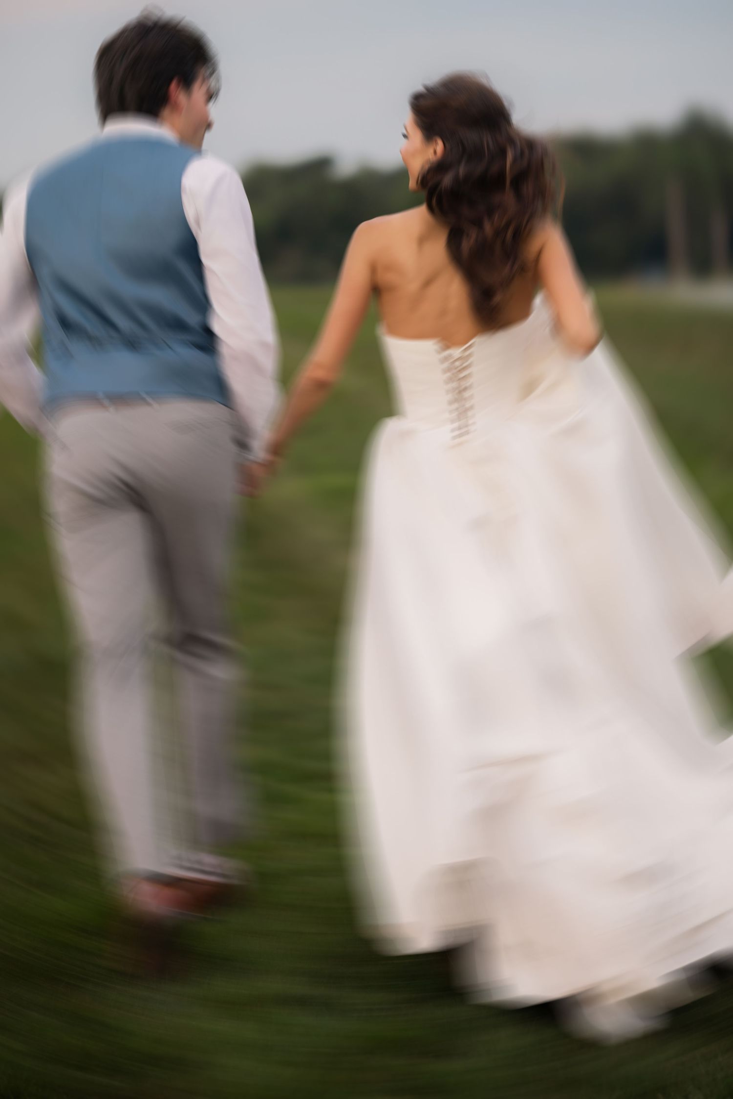 Blurred motion shot of newlyweds holding hands running through green field, bride in white gown and groom in blue vest.