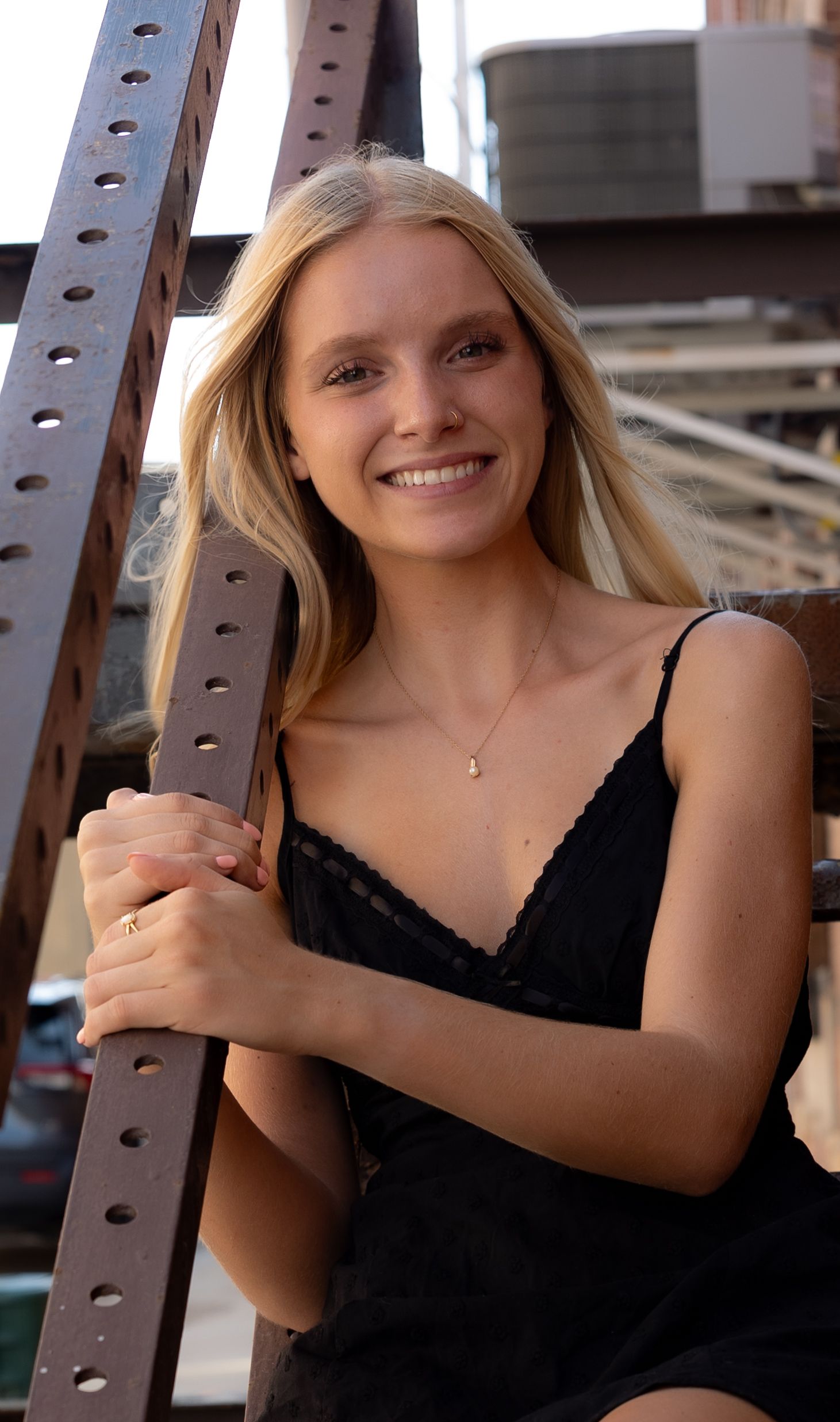Smiling blonde woman in black lace dress leaning on rusty metal beam outdoors.