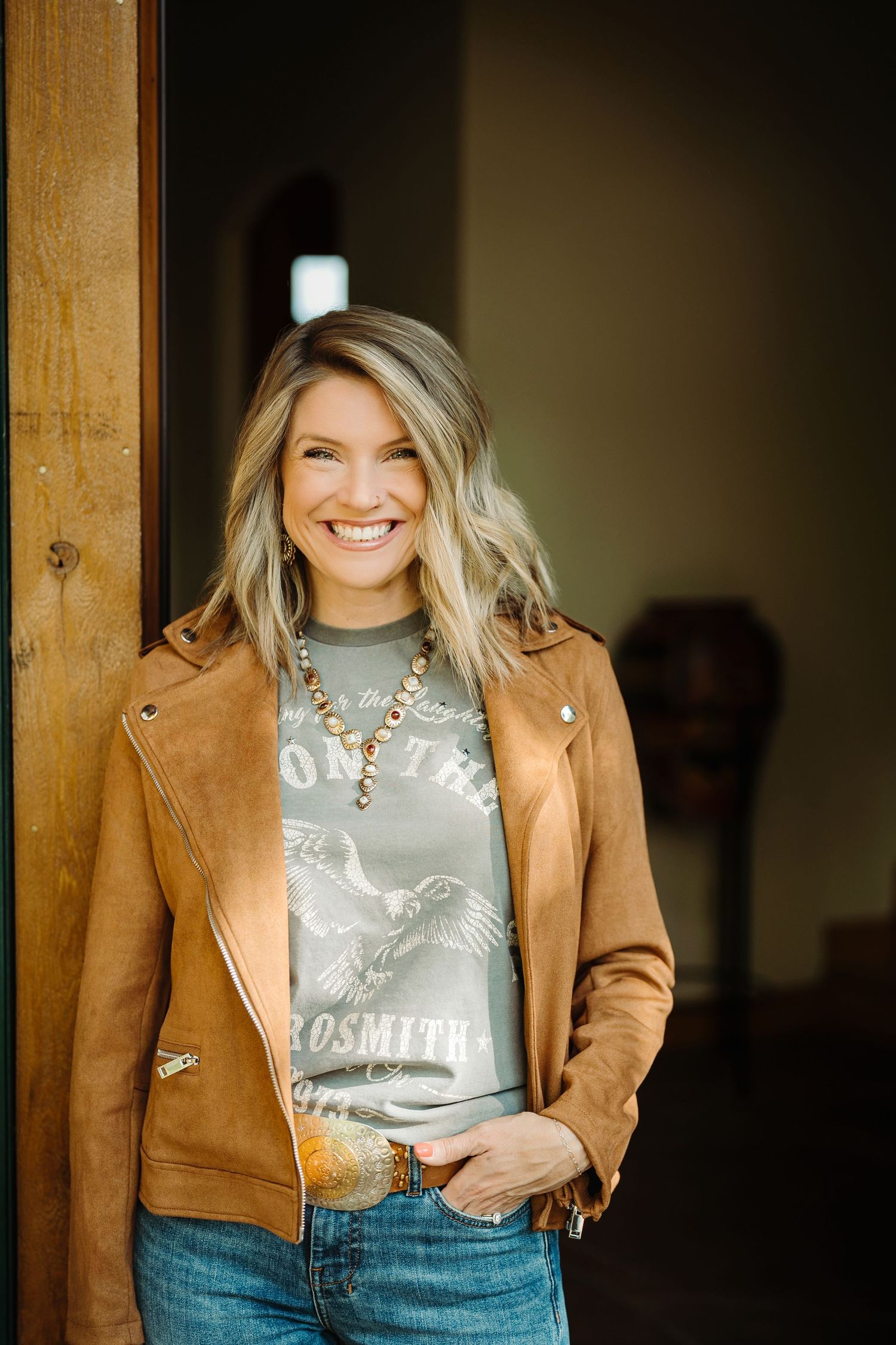 Smiling blonde woman in brown leather jacket and Aerosmith graphic tee with turquoise necklace leaning against wooden door.
