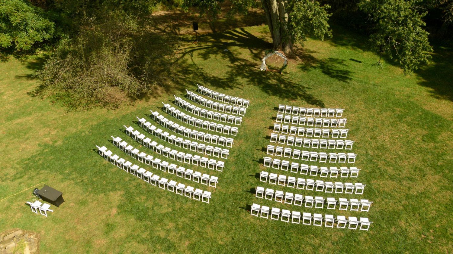 Aerial view of white wedding ceremony chairs arranged in rows on a green lawn under trees.