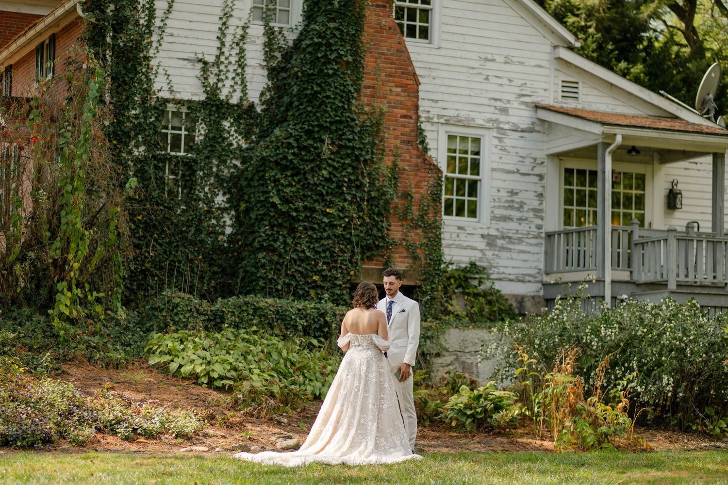 Bride and groom stand together facing an ivy-covered white farmhouse on their wedding day.