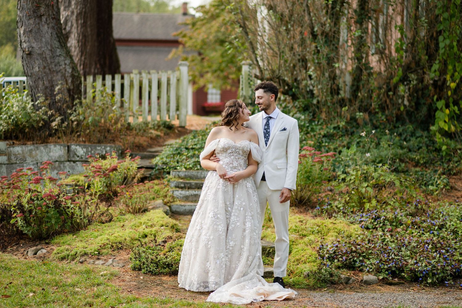 Bride and groom share a romantic moment in a lush garden with stone steps and white fence.