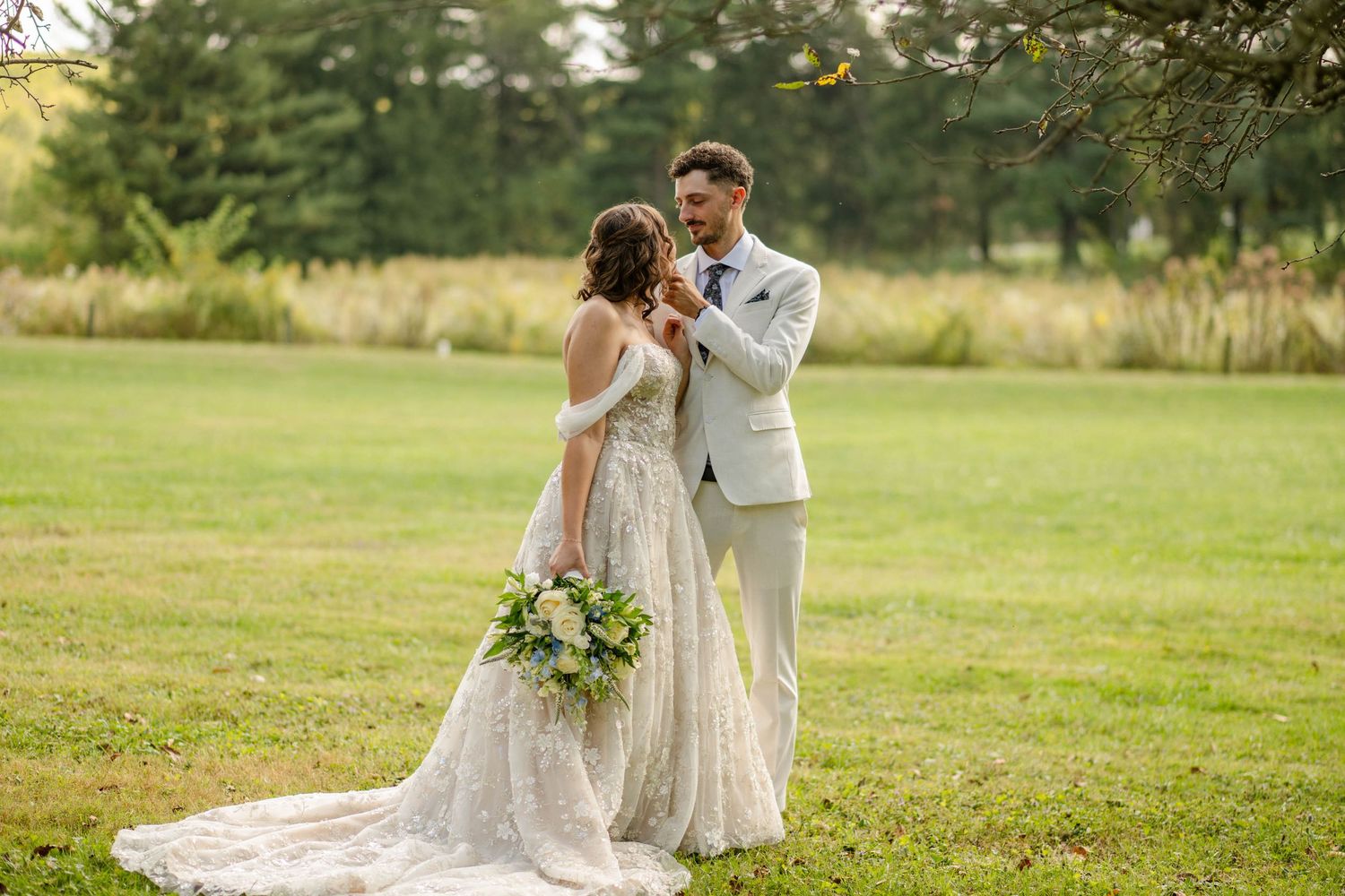 Groom tenderly holds bride holding floral bouquet while standing in an open green meadow.
