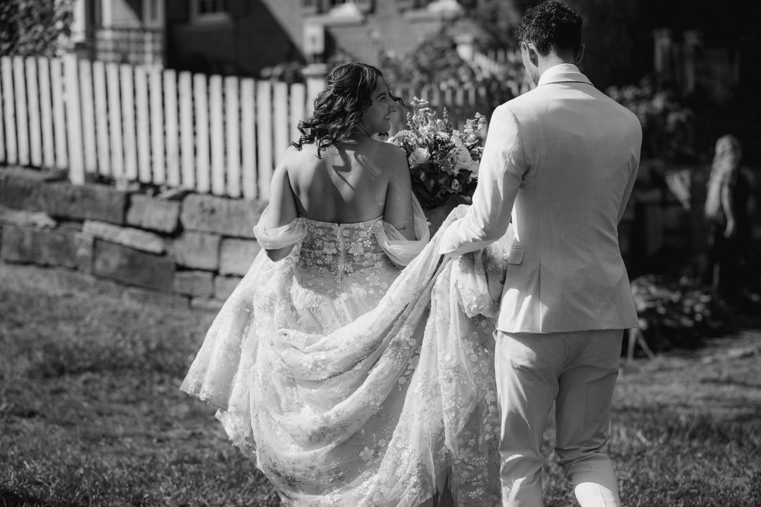 Black and white photo of groom carrying bride's lace train as they walk together outdoors.
