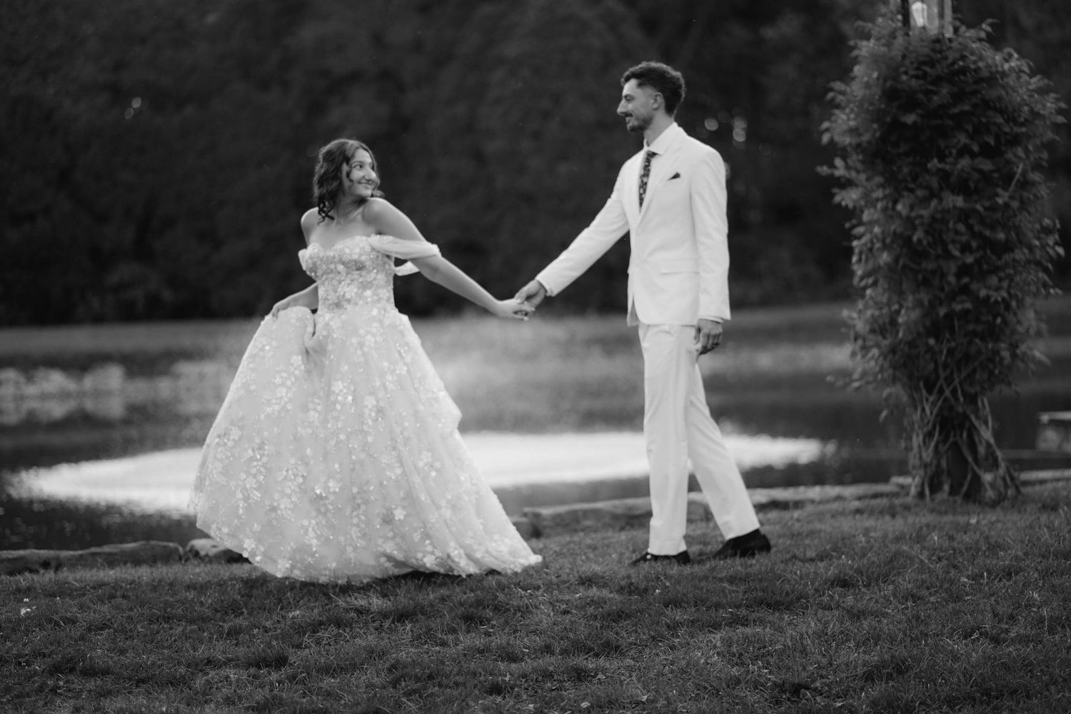 Black and white photo of bride and groom holding hands walking outdoors near a pond in wedding attire.