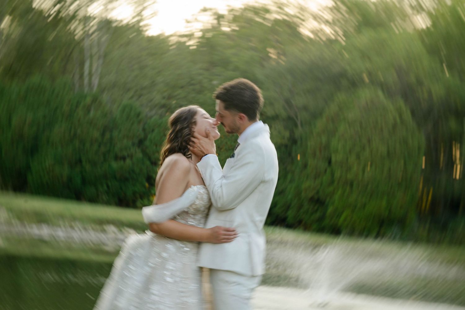 Bride and groom embrace near a sunlit pond with a dreamy motion-blur effect in nature.