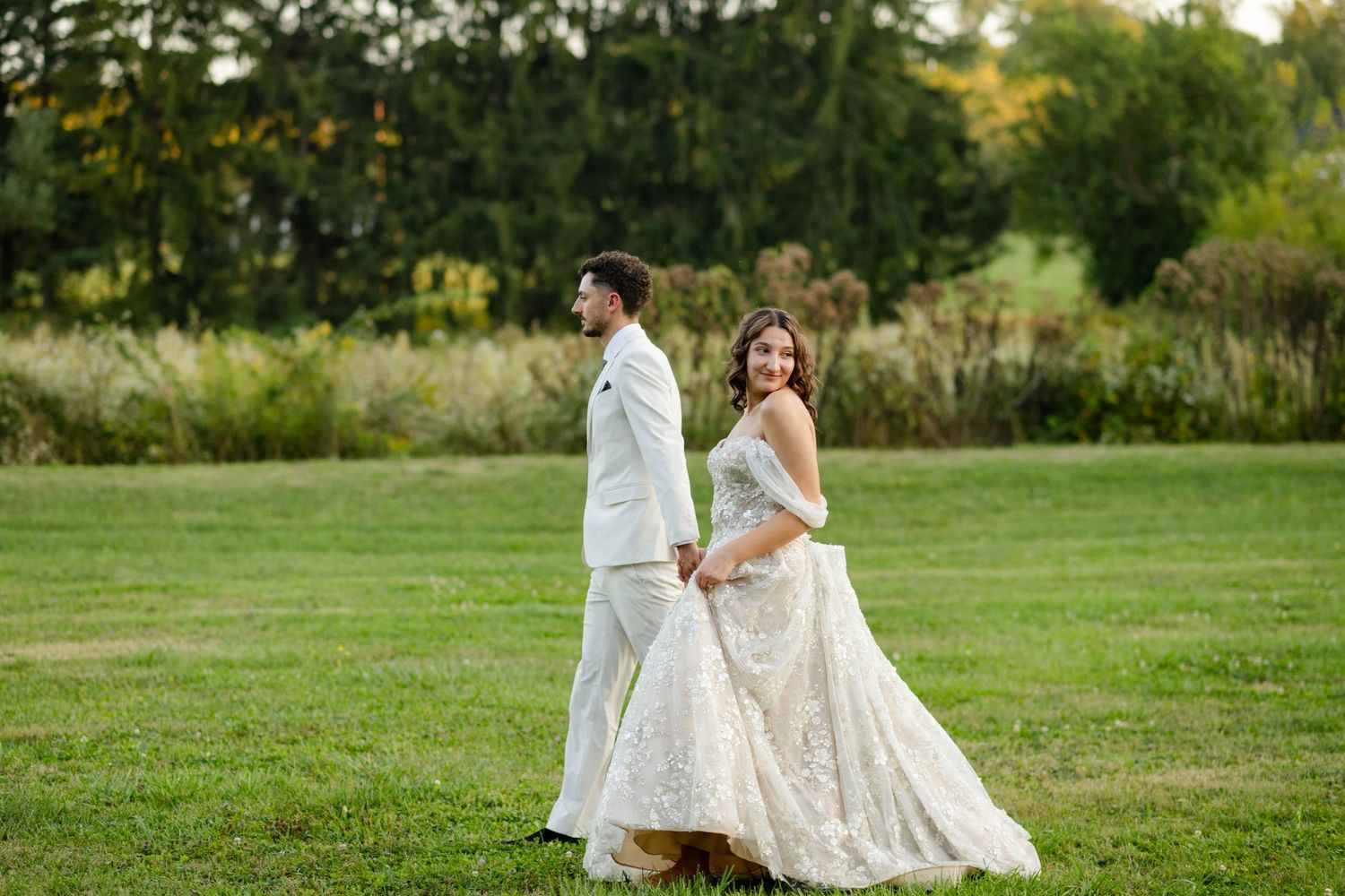 Bride glances back smiling while walking hand-in-hand with groom across a green lawn.
