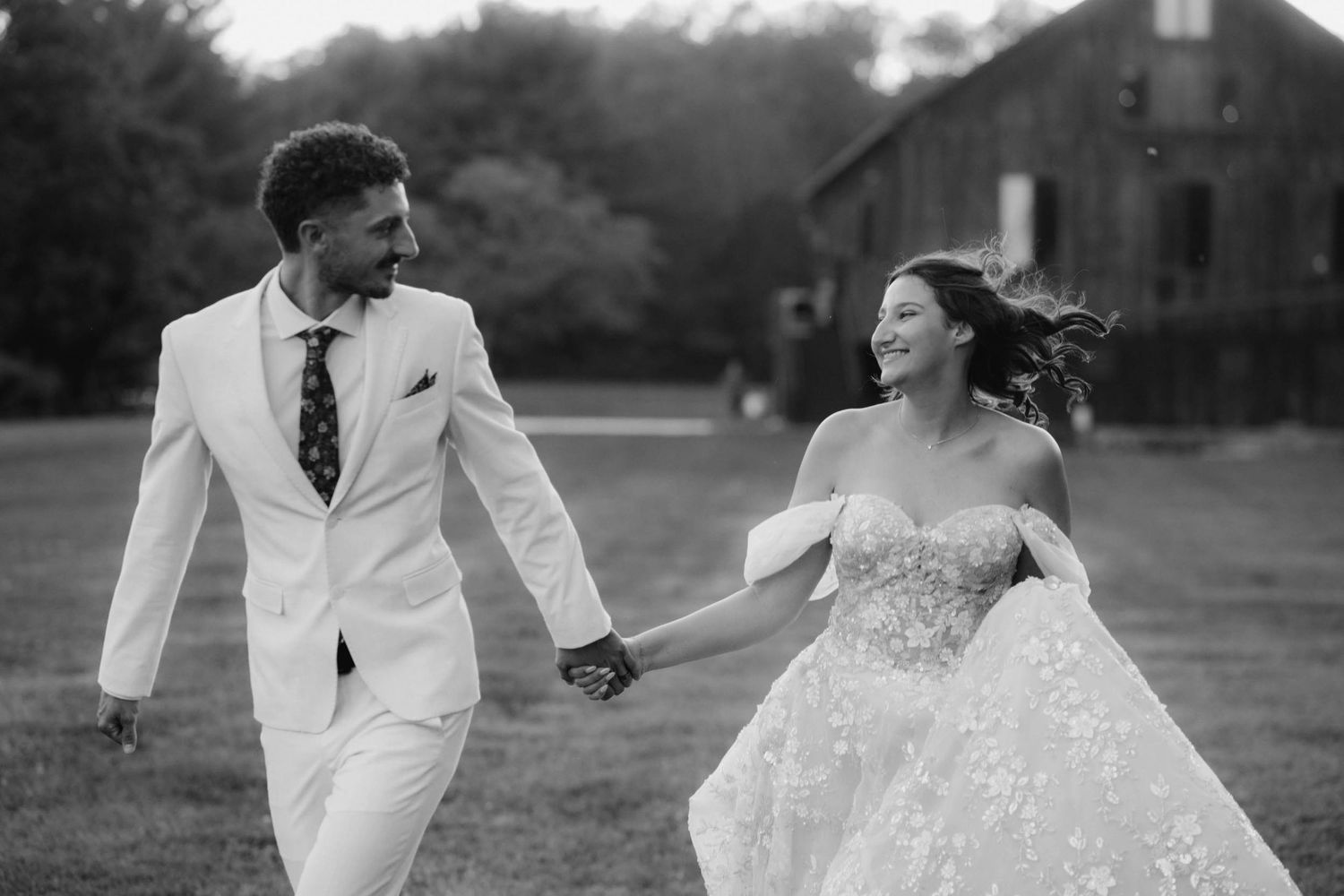 Black and white photo of bride and groom laughing and running hand in hand across a grassy field.