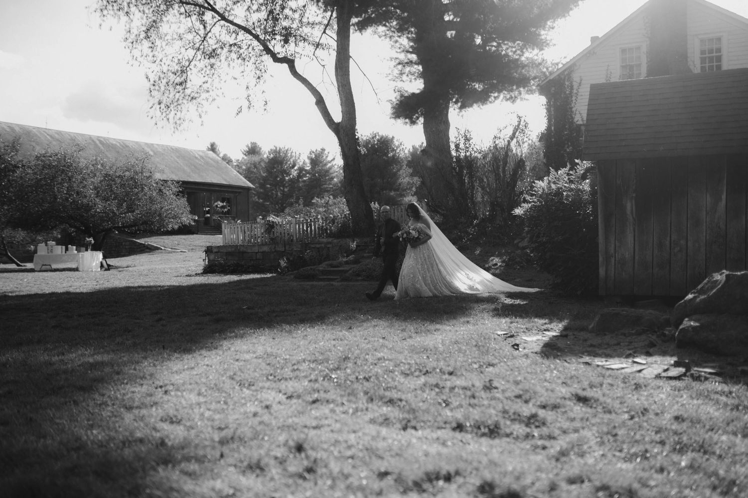 Black and white photo of bride and groom dancing near a rustic barn and trees.