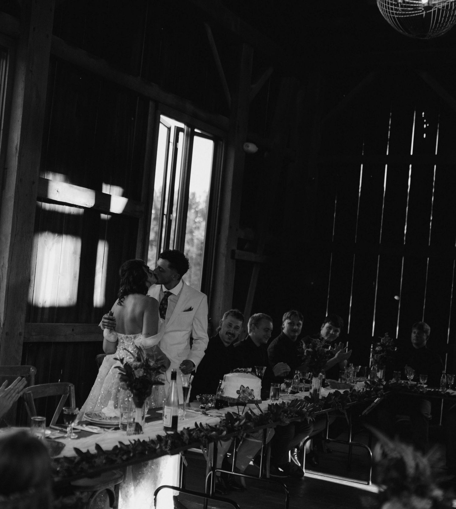 Black and white photo of couple kissing at head table during barn wedding reception.
