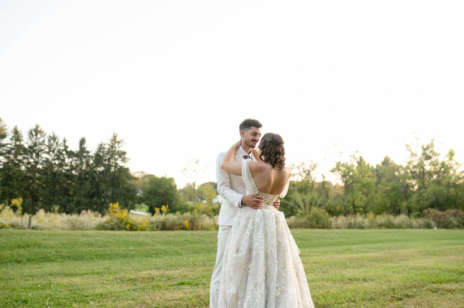 Bride and groom embrace joyfully in an open field bathed in warm sunset light.