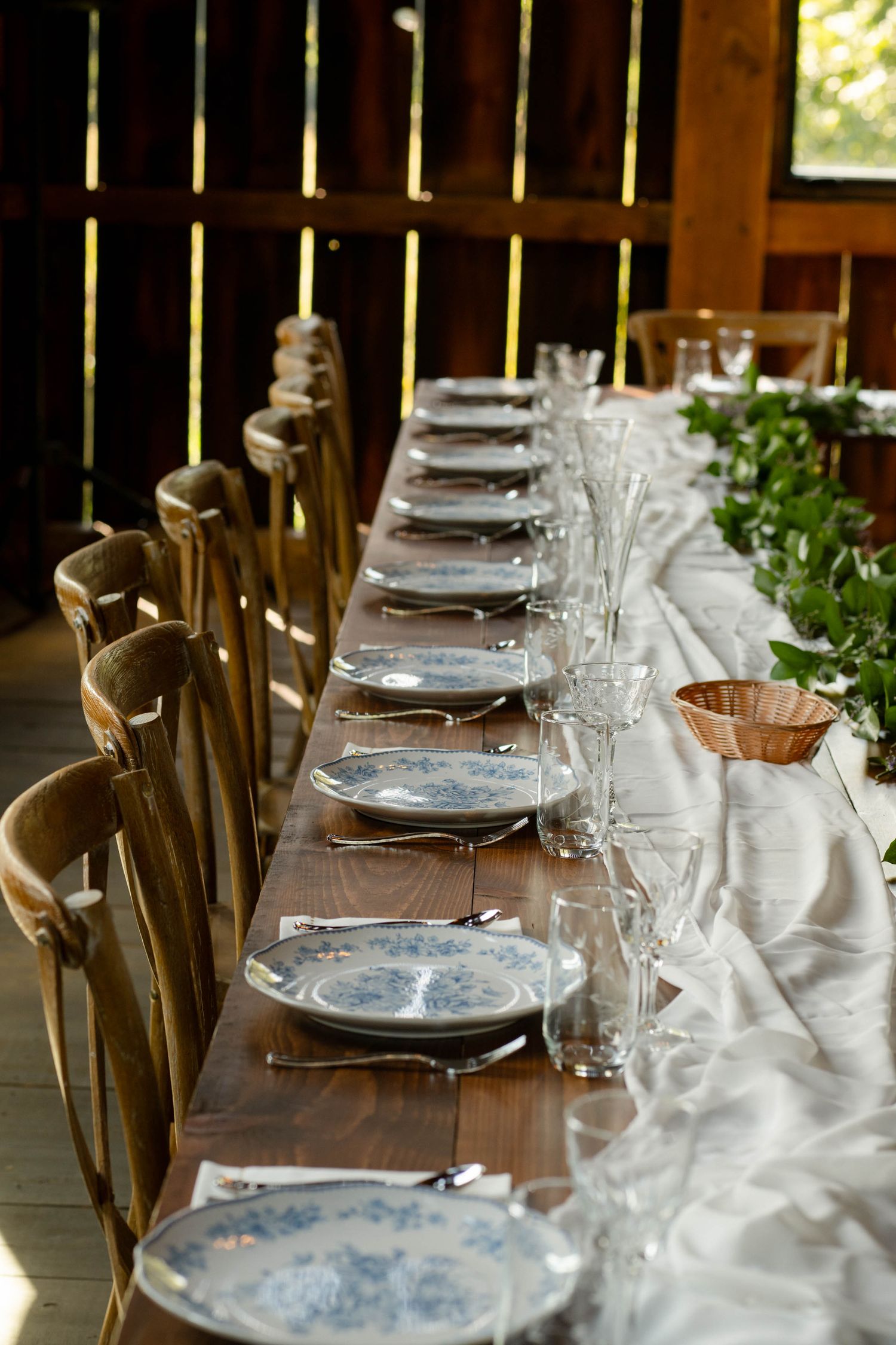 Elegant rustic barn wedding reception table set with blue floral plates, crystal glasses, and greenery.