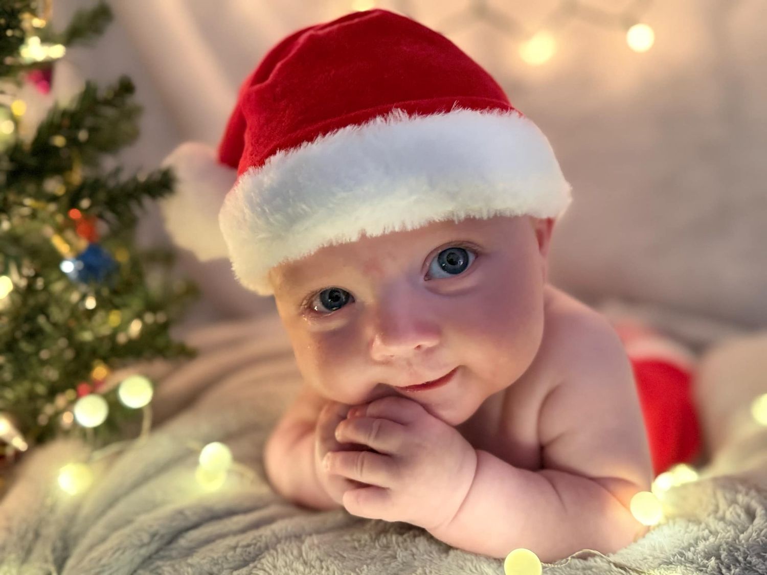 A smiling baby in a red Santa hat rests on a soft blanket near a glowing Christmas tree and twinkling holiday lights.
