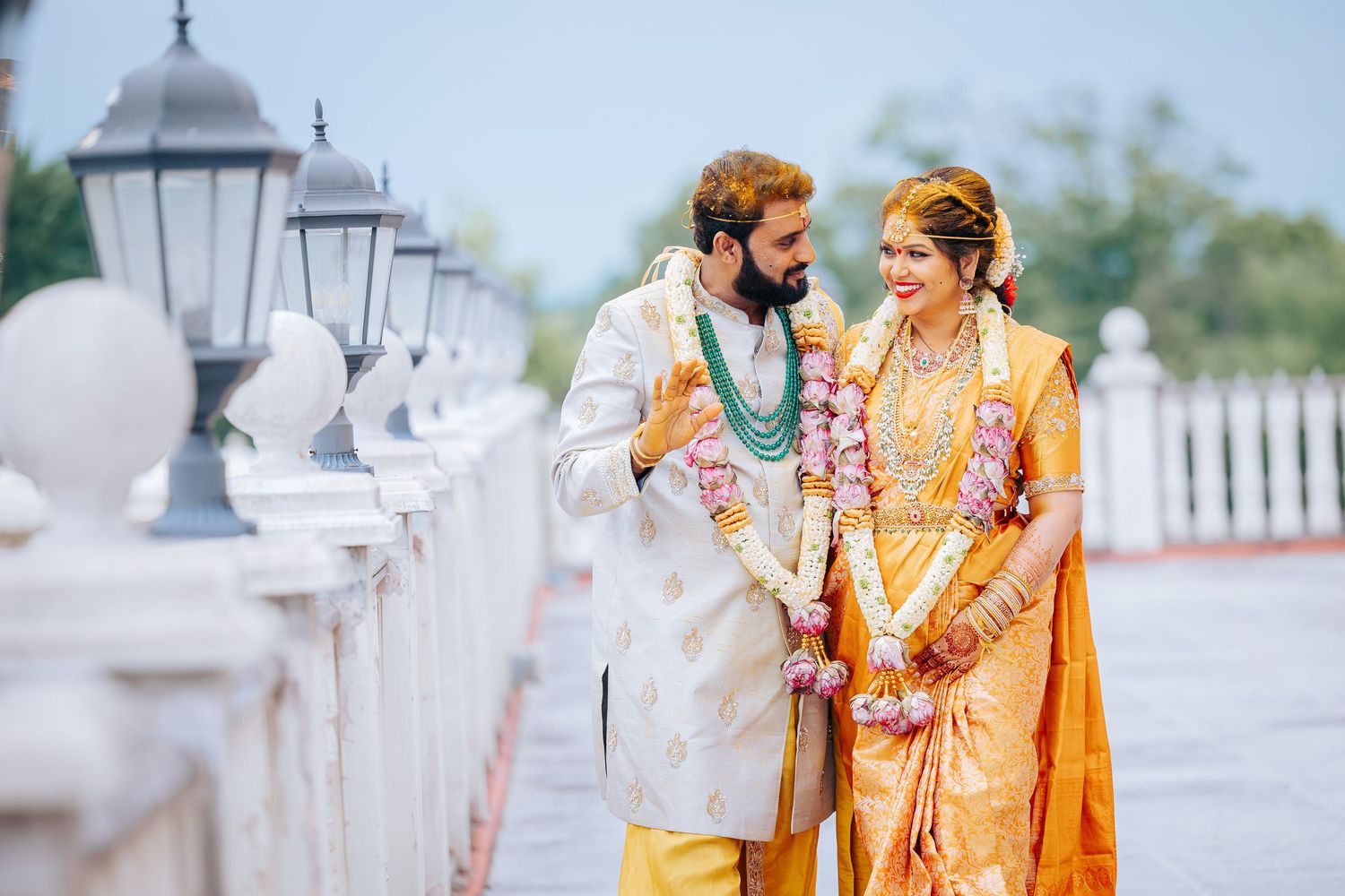 Smiling couple in traditional Indian wedding attire with flower garlands pose together on a white balustrade terrace with lanterns.