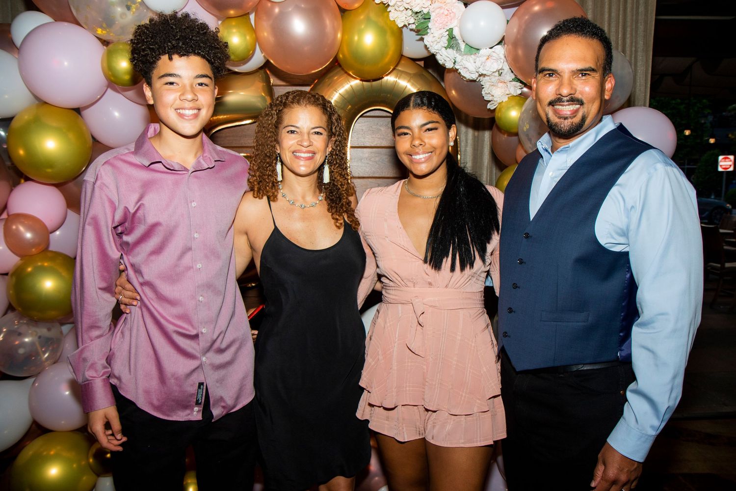 A group poses together at a celebration with gold and pink balloon decorations in the background.