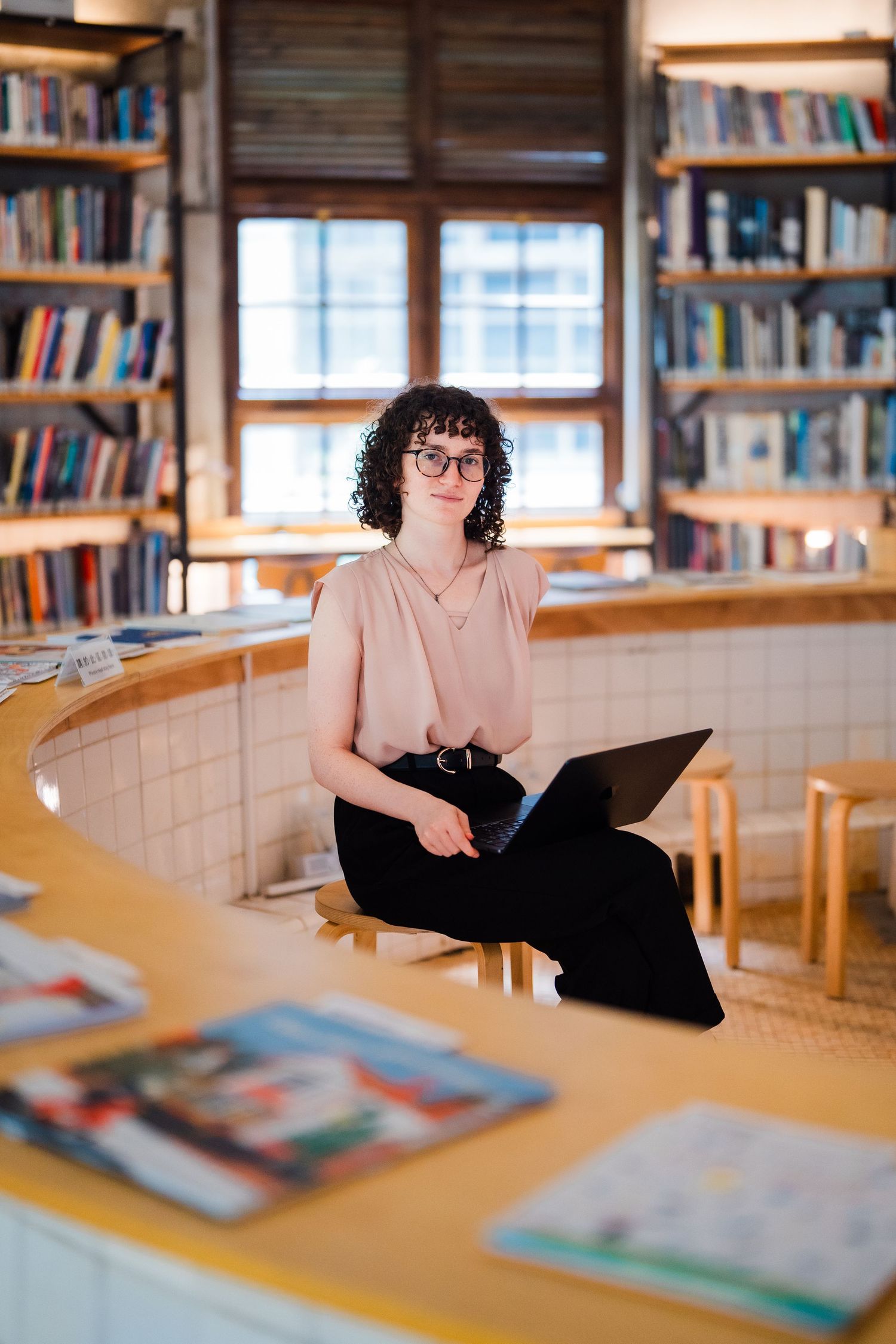 Person with laptop sits among library bookshelves near window.