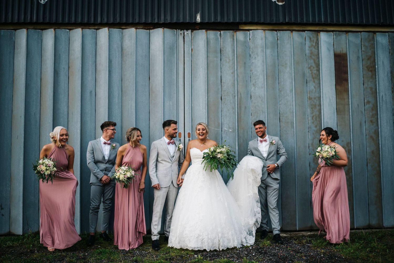 Wedding party poses against rustic metal wall with bridesmaids in pink dresses and groomsmen in gray suits.