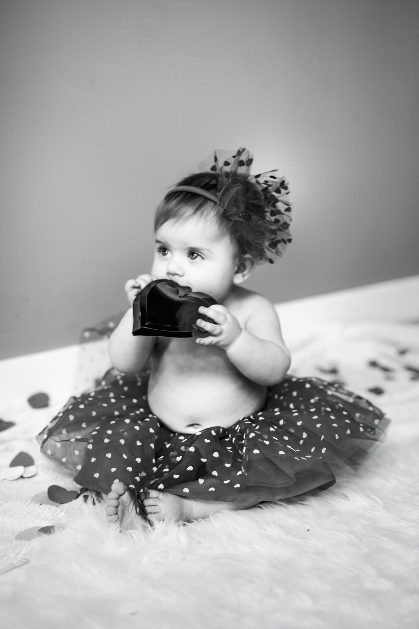Black and white photo of a baby girl in a polka dot tutu chewing on a small box.