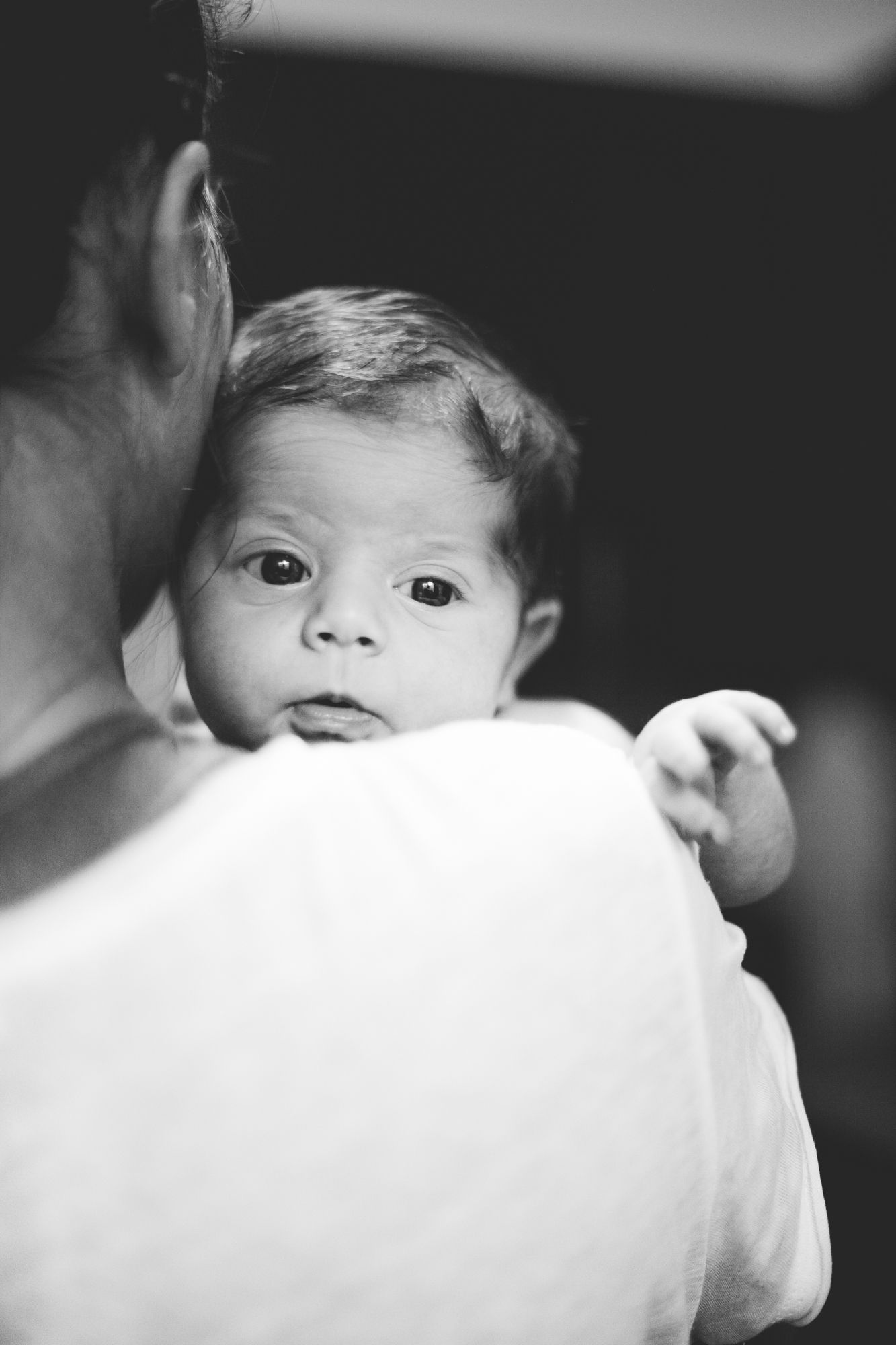 A newborn baby rests on a parent's shoulder, looking alert in a classic black and white portrait.
