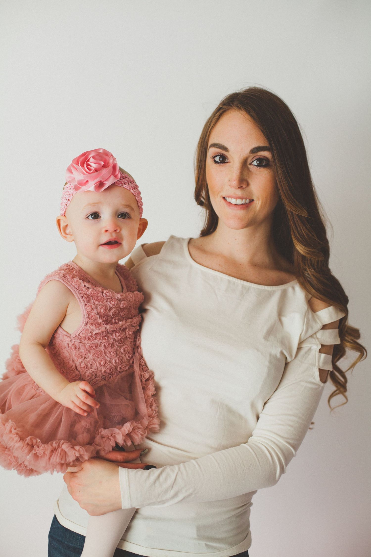 A smiling mother holds her baby girl dressed in a pink ruffled dress and floral headband against white background.