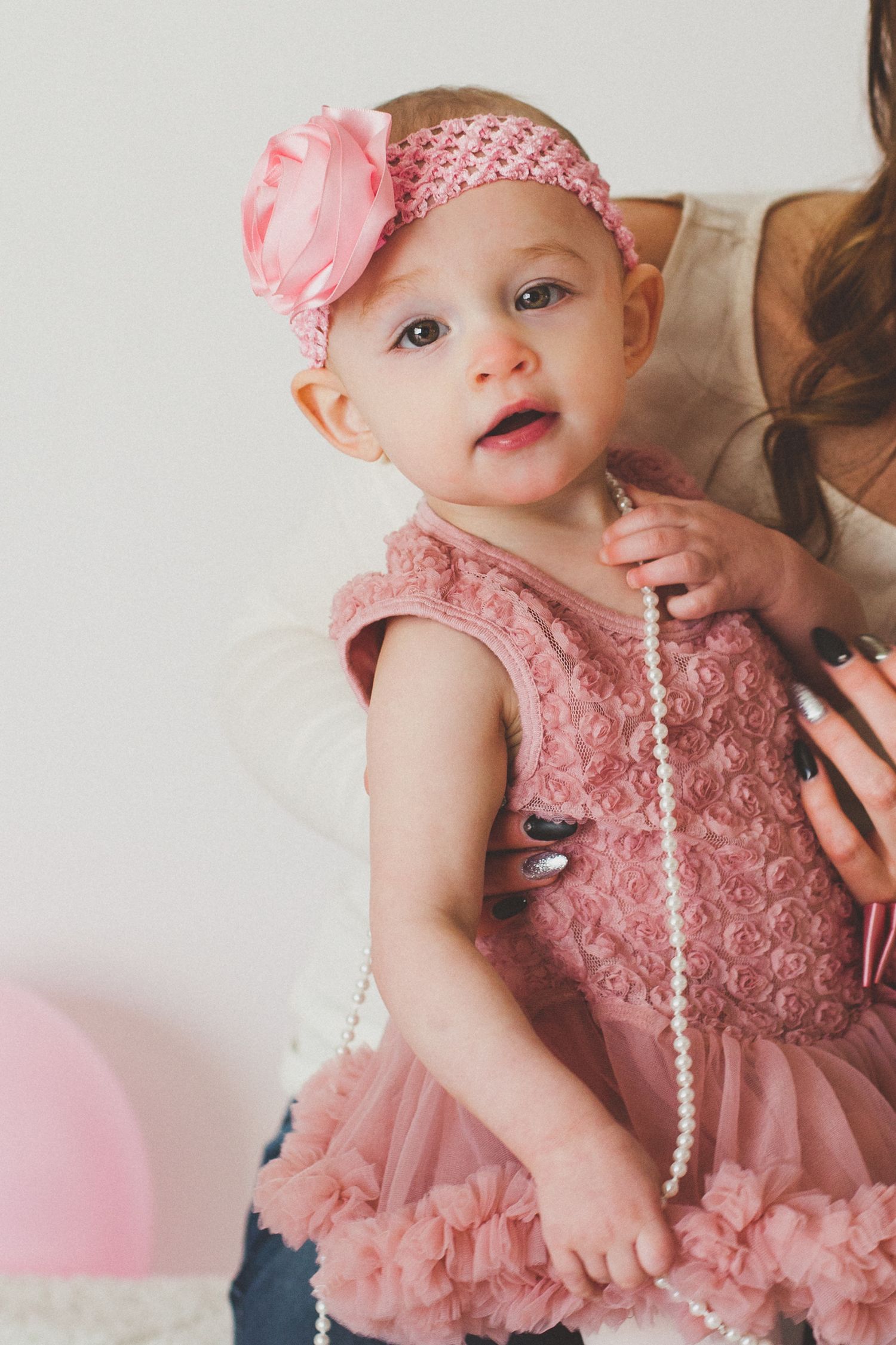 Close-up of a baby girl in pink rosette dress holding pearl necklace with mother nearby.