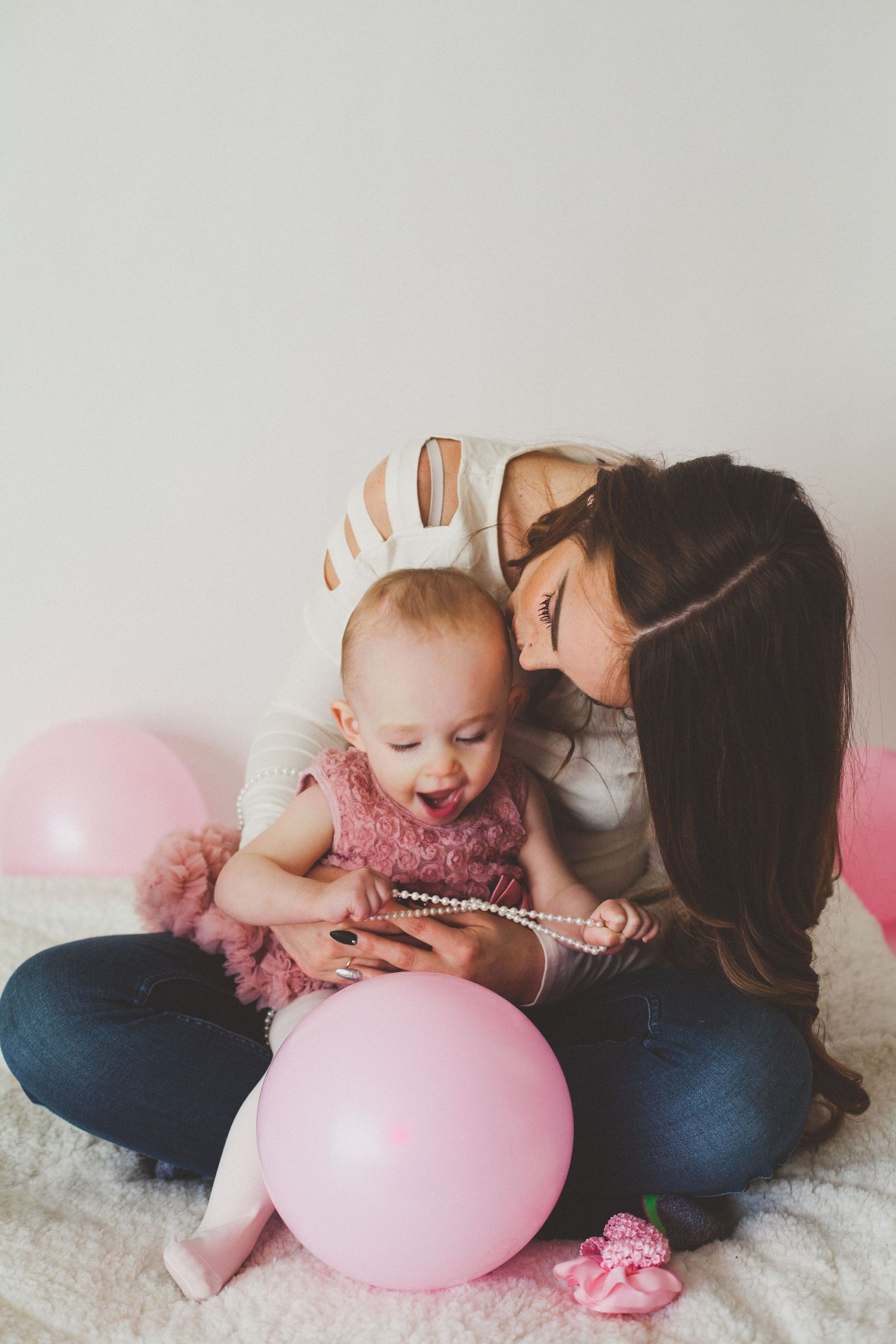 A loving mother leans over her baby girl holding pink pearl necklace with balloons nearby.