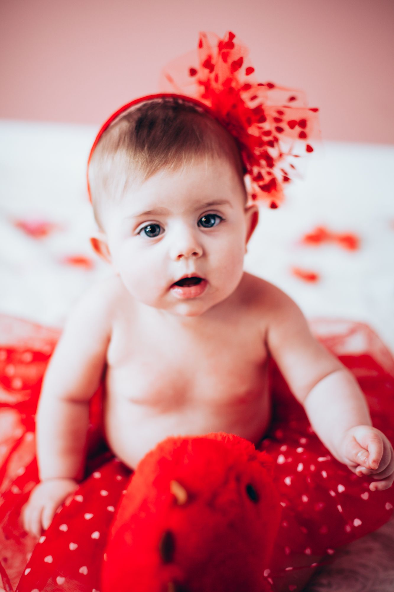 Cute blue-eyed baby wearing red bow headband and tutu skirt on white bed.