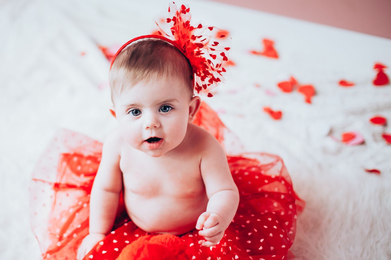 Cute baby girl in red polka dot tutu skirt and red bow headband sitting on white fur.