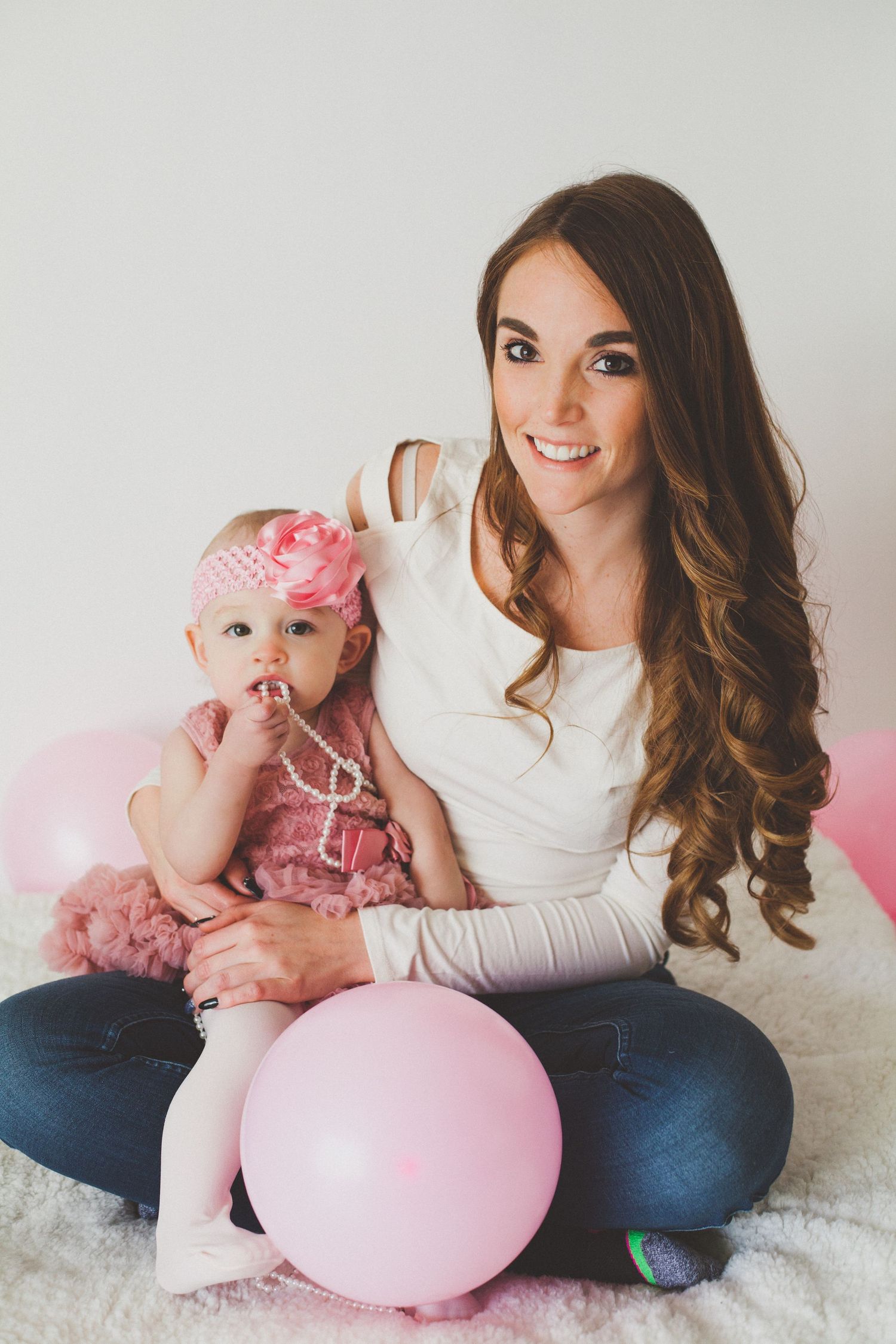 A smiling mother with long brown hair sits with her baby girl surrounded by pink balloons.