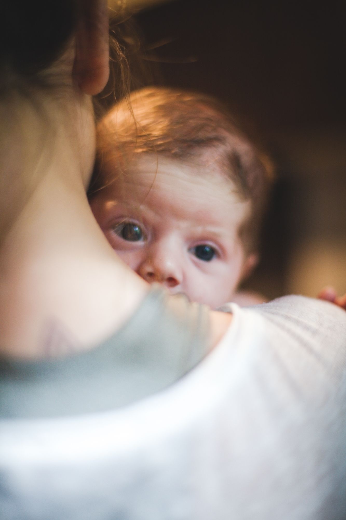 A newborn baby gazes up while being held close during breastfeeding in a warm, softly lit photo.