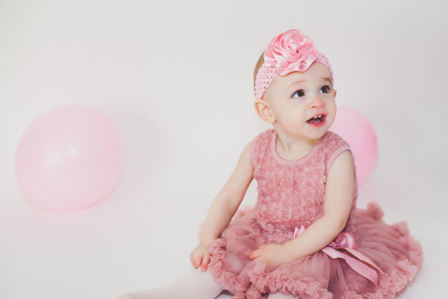 Adorable baby girl in pink ruffled dress and floral headband sitting near pink balloons.